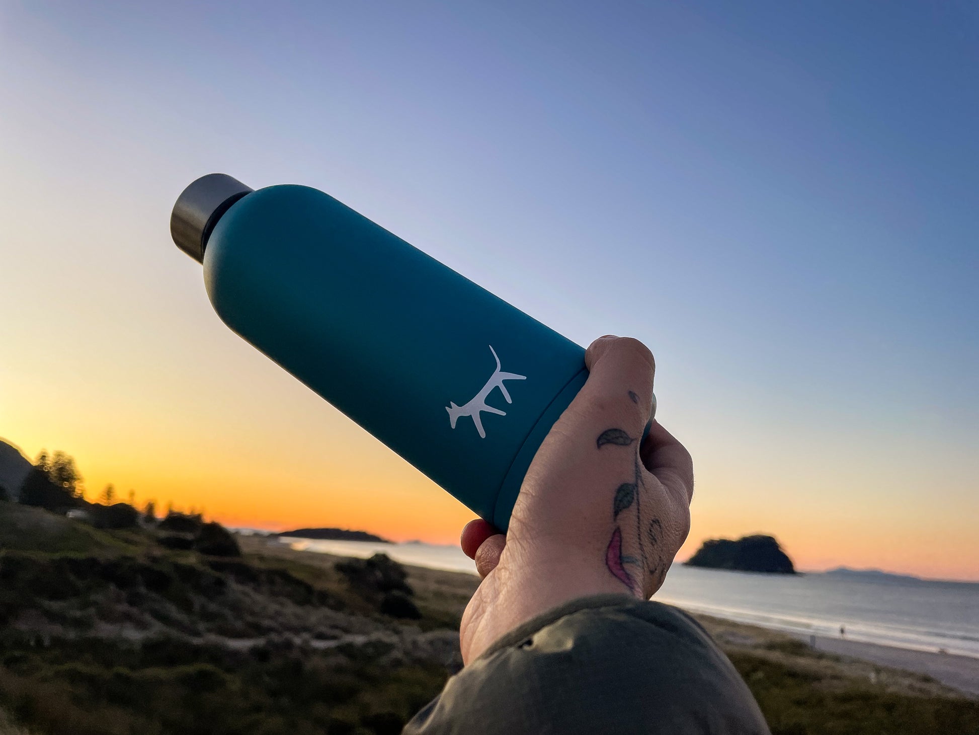 Female hand holding a stainless steel water bottle in lake colour with cream Droggo logo on the front. Beachy sunset background.