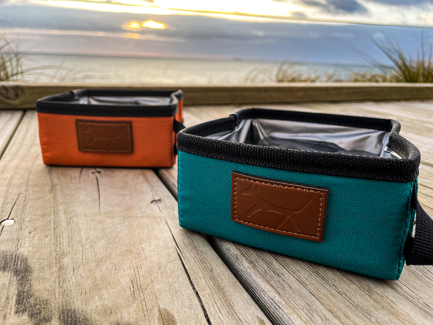 Opened dog travel bowls in rust and lake colours with leather Droggo logo on the front right beside each other. Both travel bowls are on top a wooden deck at the beach. Sunset scene on the background.