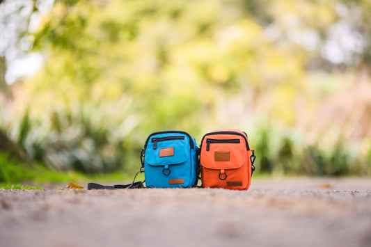 Dog walking bag in lake and rust colour with leather Droggo logo on the front right beside each other. Both bags are laying on a reserve pathwalk.