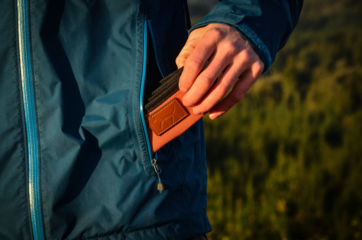 Male hand putting a dog travel bowl in rust colour with leather Droggo logo on the front in his blue rain jacket pocket. Pine forest blurred background.