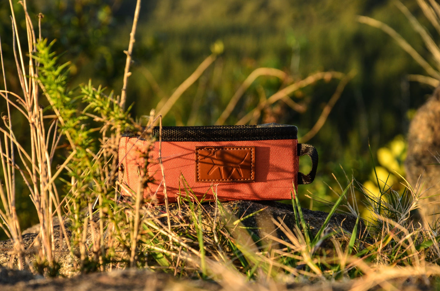 Oxford dog travel bowl in rust colour and leather Droggo logo in the sunlight on the ground of track with some shrub around it.